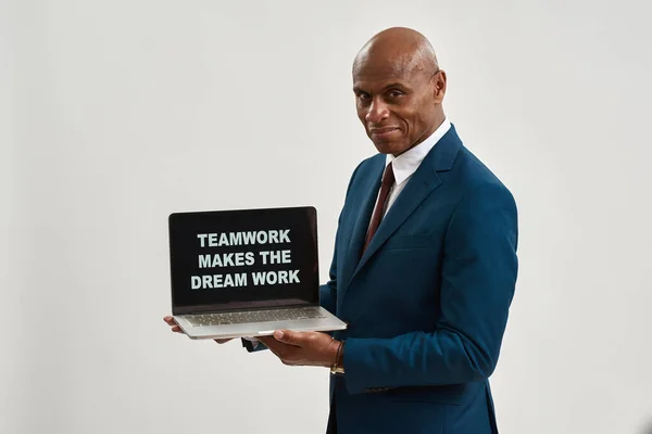 Smiling black businessman holding laptop computer with teamwork makes the dream work slogan. Adult man wear formal wear. Modern successful male lifestyle. White background. Studio shoot. Copy space
