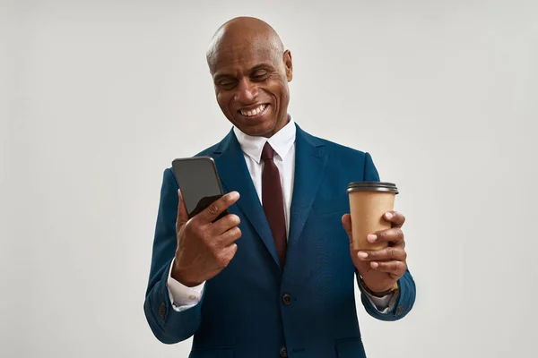 Smiling black entrepreneur with coffee watching smartphone. Bald adult man wearing formal wear. Concept of modern successful male lifestyle. Isolated on white background. Studio shoot. Copy space