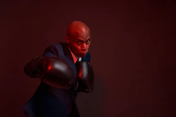 Confident black businessman boxing on dark red background. Adult man wearing formal wear and boxing gloves. Modern successful male lifestyle. Business goal. Leadership. Studio shoot. Copy space
