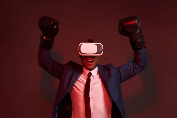 Excited african american businessman wearing boxing gloves and virtual reality glasses celebrating win or success. Concept of business goal. Leadership. Dark red background. Studio shoot. Copy space