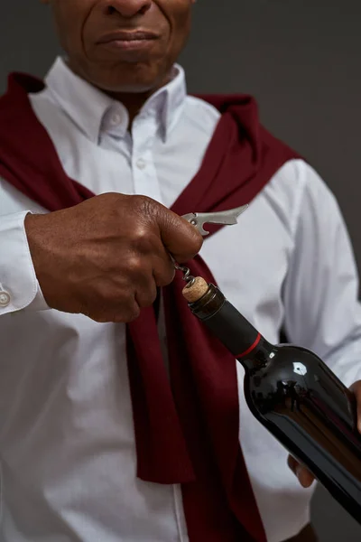 Partial and obscure face of black winemaker or sommelier opening wine bottle with corkscrew. Adult successful male entrepreneur. Viticulture and winemaking. Isolated on grey background. Studio shoot