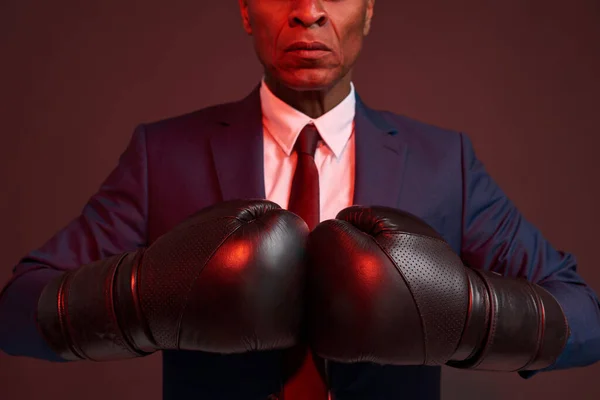 Obscure face of adult serious black businessman wearing formal suit and boxing gloves. Modern successful male lifestyle. Concept of business goal. Leadership. Dark red background. Studio shoot