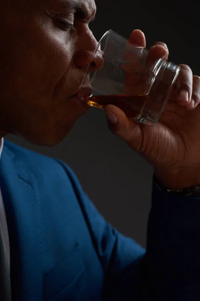 Cropped of black company leader with closed eyes drinking cognac or brandy from glass. Adult man wearing formal wear. Modern successful male lifestyle. Dark red background. Studio shoot. Copy space