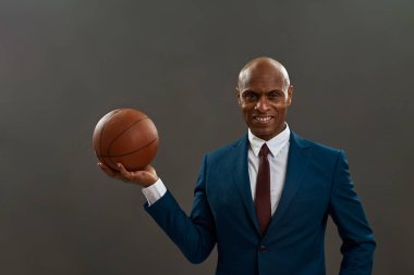Basketbol topu tutan gülümseyen siyah girişimci. Yetişkin adam resmi kıyafet giyiyor. Modern başarılı erkek yaşam tarzı. İş hedefi kavramı. Gri arka planda izole edilmiş. Stüdyo çekimi. Boşluğu kopyala