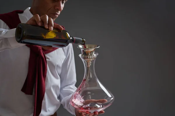 Obscure face of focused adult african american winemaker or sommelier pour red wine in decanter from bottle. Wine tasting. Viticulture and winemaking. Isolated on grey background in studio. Copy space