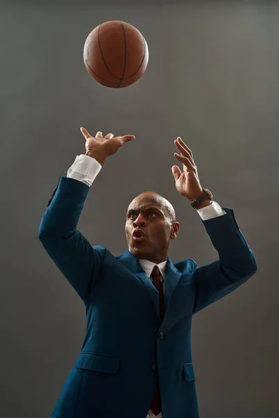 Concentrated black entrepreneur or ceo manager throwing basketball ball. Bald adult man wearing formal wear. Modern successful male lifestyle. Concept of business goal. Grey background. Studio shoot