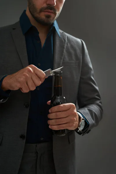 Obscure face of brewer entrepreneur opening beer bottle with corkscrew. Bearded man wearing formal wear. Concept of brewing business. Modern successful male lifestyle. Grey background. Studio shoot