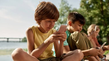 Bulanık arkadaşları olan ve nehir kıyısında akıllı telefonları kullanan seçici bir erkek çocuğu. Alfa neslinin güzel çocukları. Gadget bağımlılığı. Çocukluk yaşam tarzı. Güneşli yaz günü