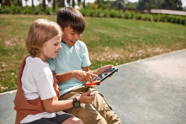 Çok ırklı bir çocuk açık havada beyaz erkek çocuk arkadaşı için oyun konsolunu işaret ediyor. Dijital nesil alfa 'nın konsantre çocukları. Modern çocukluk tarzı. Gadget bağımlılığı. Güneşli bir gün