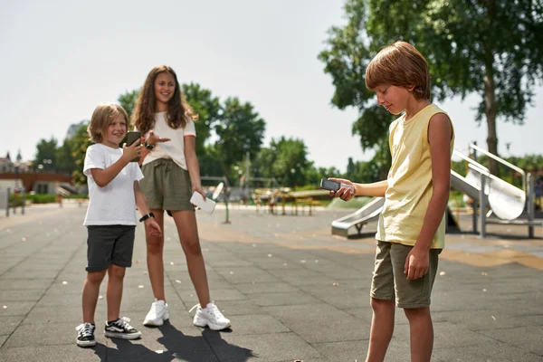 Children with modern smartphones laughing from embarrassed boy with push-button mobile phone on playground. Digital generation alpha. Gadget addiction. Childhood lifestyle. Kid bullying. Sunny day