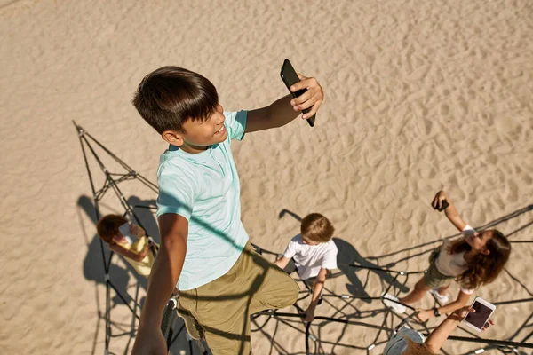Top view of multiethnic children searching mobile connection on climbing net on playground outdoors. Boys and girls of modern generation alpha. Gadget addiction. Childhood lifestyle. Sunny day