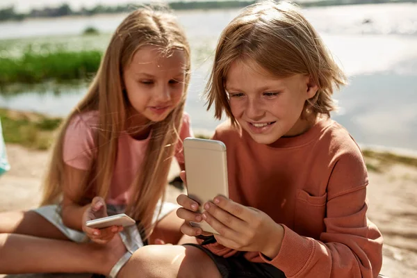 Girl looking at her boy friend watching mobile phone on blurred sandy coast outdoors. Beautiful caucasian kids of generation alpha. Gadget addiction. Childhood lifestyle. Friendship. Warm summer day