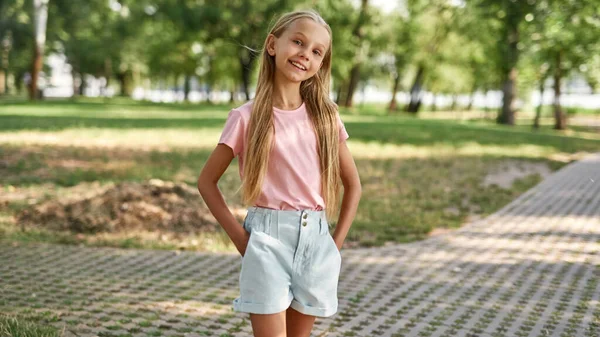 在绿油油的公园里看着镜头 双手插在口袋里笑着的欧洲姑娘 漂亮的金发女宝宝阿尔法一代穿着T恤和短裤 童年的生活方式阳光灿烂的夏日 — 图库照片