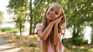 Uyurken gülümseyen beyaz kız bulanık yeşil parktaki kameraya bakıyor. Alfa neslinin güzel sarışın kızı tişört giyiyor. Çocukluk yaşam tarzı kavramı. Güneşli yaz günü