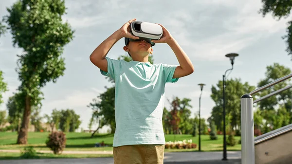 Focused boy using Virtual reality headset outdoors. Dark-haired male child of digital generation alpha. Concept of modern childhood lifestyle. Gadget addiction. Green park in sunny summer day