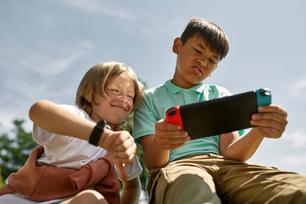 Smiling boy with thumb down looking at dissatisfied multiracial male child friend playing video game on game console outdoors. Digital generation alpha. Modern childhood lifestyle. Gadget addiction