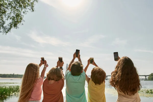 孩子们在河边用智能手机拍摄蓝天阳光的背景图片 一代的男孩和女孩阿尔法 小工具上瘾 童年的生活方式温暖的夏日 — 图库照片