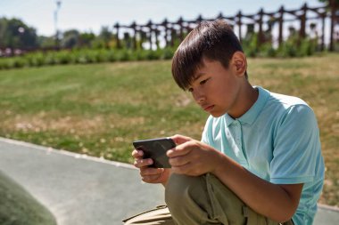 Açık havada akıllı telefondan video oyunu oynayan çok ırklı bir çocuk. Dijital nesil Alfa 'nın koyu saçlı erkek çocuğu. Modern çocukluk yaşam tarzı kavramı. Gadget bağımlılığı. Güneşli yaz günü