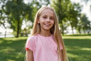 Açık havada bulanık yeşil parkta kameraya bakan gülümseyen beyaz bir kızın kısmi görüntüsü. Alfa neslinin güzel sarışın kızı tişört giyiyor. Çocukluk yaşam tarzı. Güneşli yaz günü