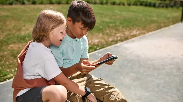 Dışarıdaki oyun konsolunda oyun oynayan çoklu etnik kökenli erkek çocuk arkadaşına bakan gülümseyen çocuk. Dijital nesil alfa çocukları. Modern çocukluk tarzı. Gadget bağımlılığı. Güneşli bir gün