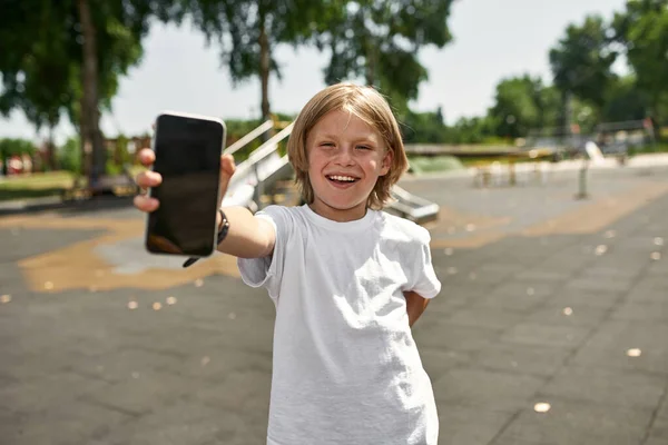 Leende Kaukasiska Pojke Visar Smartphone Och Tittar Kameran Suddig Lekplats — Stockfoto