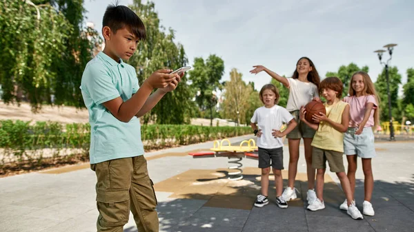 Skupina Kavkazských Dětí Volá Osamělého Multiraciálního Chlapce Pomocí Smartphonu Hřišti — Stock fotografie