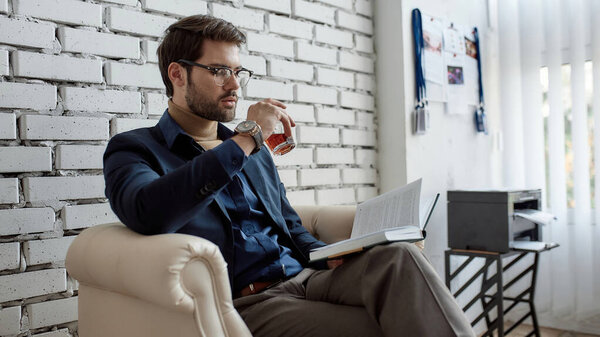 Сосредоточенный кавказский IT-инвестор или продавец читает книги и пьет коньяк из стекла в кресле в офисе. Бизнес-стратегия, развитие, продвижение, продажа и маркетинг высокотехнологичной компании