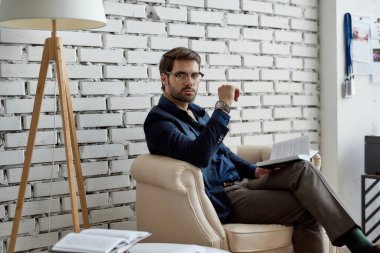 Ofisteki koltukta kitap okurken kameraya bakan kendinden emin beyaz bilişim satıcısı. Yüksek teknoloji şirketinin iş stratejisi, geliştirilmesi, tanıtımı, satışı ve pazarlanması. Başarılı erkek yaşam tarzı.