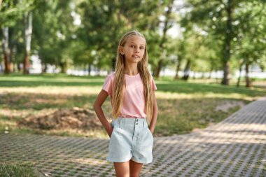 Elleri ceplerinde gülümseyen beyaz kız bulanık yeşil parktaki kameraya bakıyor. Tişört ve şort giyen alfa neslinin tatlı sarışın kızı. Çocukluk yaşam tarzı. Güneşli yaz günü