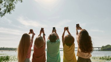 Bir grup çocuğun nehir kıyısındaki akıllı telefonlarıyla mavi gökyüzünde güneşin fotoğrafını çekerken. Alfa neslinin kızları ve erkekleri. Gadget bağımlılığı. Çocukluk yaşam tarzı. Yaz gündüz