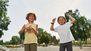 Arkadaşının yanında dans eden çocuk dışarıda, bulanık oyun alanında kulaklıkla müzik dinliyor. Dijital nesil alfa 'nın beyaz erkek çocukları. Gadget bağımlılığı. Çocukluk yaşam tarzı. Arkadaşlık. Güneşli bir gün