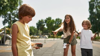 Modern akıllı telefonları olan çocuklar dışarıda oyun parkında düğmeli telefonlarıyla gülerek gücenmiş çocuğa işaret ediyorlar. Dijital nesil alfa. Gadget bağımlılığı. Çocukluk yaşam tarzı. Çocuk zorbalığı
