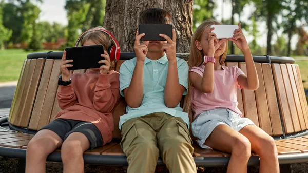 一群孩子坐在户外的木制长椅上 一边使用和浏览智能手机 一代的男孩和女孩阿尔法 小工具上瘾 童年的生活方式阳光灿烂的夏日 — 图库照片