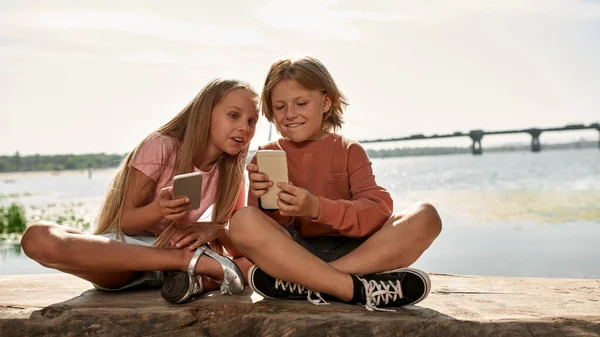 Soustředěný Chlapec Překvapená Dívka Sledující Smartphone Log Břehu Řeky Venku — Stock fotografie