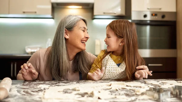 小さな女の子と顔に小麦粉と高齢者の女性がお互いを見て笑っている 祖母は孫に家庭のキッチンでクッキーを調理することを教えている 家族関係と楽しい時間を一緒に — ストック写真
