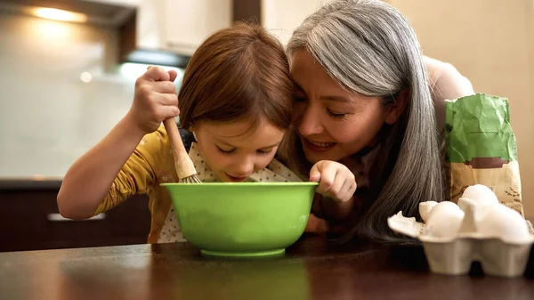 Caucasian little girl mixing dough in bowl while grandmother looking this. Senior woman teaching granddaughter cooking. Family education and spend time together. People at table at home kitchen