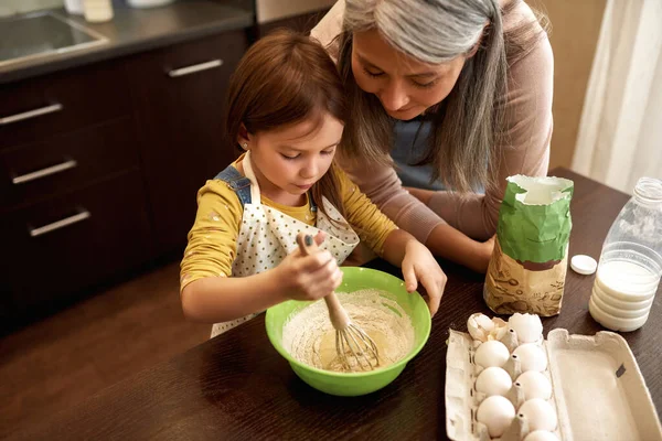 European little girl mixing dough in bowl while grandmother looking this. Senior woman teach granddaughter cooking. Concept of family education and spend time together. People at table at home kitchen