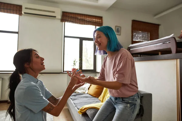 Girl standing on knee and presenting wedding ring to her pleased girlfriend at spacious home. Young caucasian lesbain couple. Wedding proposal and marriage. Romantic homosexual relationship