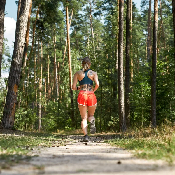 Yeşil ormanda koşan ya da koşan genç bir sporcunun arka plan görüntüsü. Spor kıyafetleri giyen, çekici, sarışın, zayıf kadın. Modern sağlıklı yaşam tarzı kavramı. Güneşli yaz günü