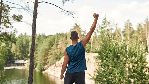 Back view of successful sportsman with hand in air celebrating his win or triumph on top stone hill on river or lake coast in forest. Man wearing sportswear. Achieving of sports goal. Warm summer day