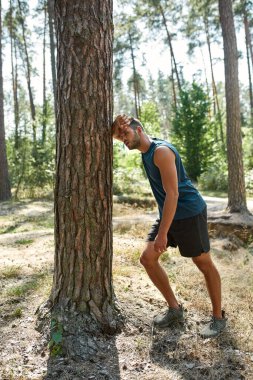 Bitkin düşmüş beyaz tenli sporcu ağaca dokunuyor ve yeşil ormanda koştuktan sonra dinleniyor. Yakışıklı, sakallı, siyah saçlı adam spor giyer. Modern sağlıklı yaşam tarzı kavramı. Güneşli yaz günü