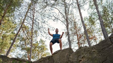 Genç beyaz tenli başarılı sporcunun zaferini ya da ormandaki en tepedeki zaferini kutlaması. Erkek adam spor giyer. Spor hedefine ulaşmak için. Modern sağlıklı yaşam tarzı. Sıcak yaz günü