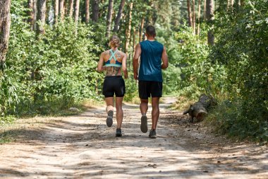 Yeşil Orman 'da koşan genç bir çiftin arka görüntüsü. Dövmeleri olan güzel, sarışın, zayıf bir kadın ve spor giyimli yakışıklı bir adam. Modern sağlıklı yaşam tarzı kavramı. Güneşli yaz günü