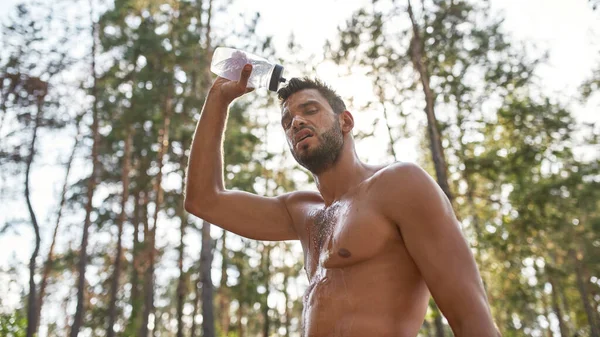 Exhausted young caucasian sportsman pouring water on himself from bottle in blurred forest. Handsome bearded man with naked torso and closed eyes. Concept of modern healthy lifestyle. Warm summer day