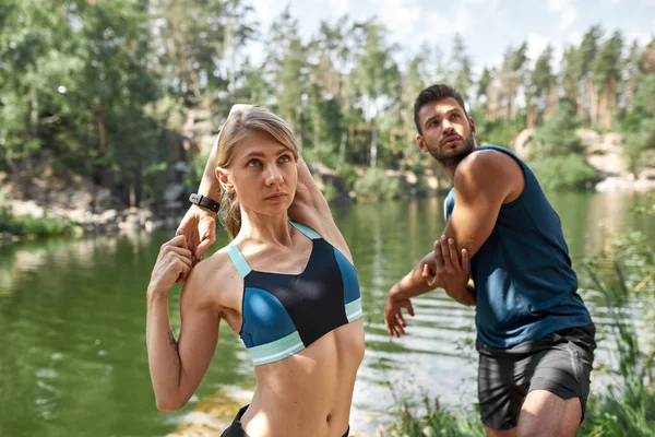 Young caucasian sports couple stretching on river or lake coast. Attractive blonde slim woman and handsome bearded man wearing sportswear. Concept of modern healthy lifestyle. Sunny summer day