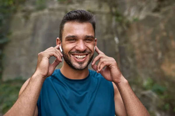 Portrait of smiling young caucasian sportsman choosing music in wireless earphones outdoors. Handsome bearded man wearing sportswear looking at camera. Concept of modern healthy lifestyle