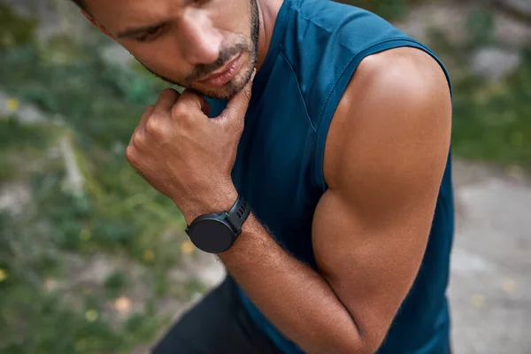 Obscure face and partial of young caucasian sportsman thinking about something outdoors. Handsome athletic man wearing sportswear and smartwatch. Concept of modern healthy lifestyle. Warm summer day