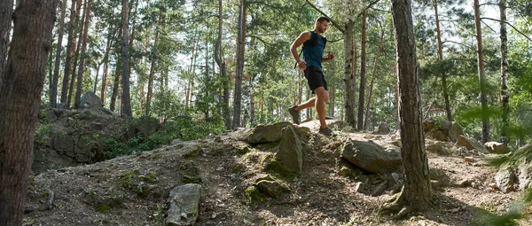Young caucasian sportsman running or jogging on hill in green forest. Handsome bearded dark-haired athletic man wearing sportswear. Concept of modern healthy lifestyle. Sunny summer daytime