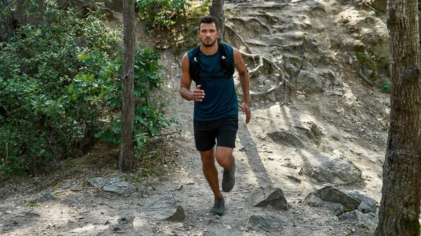 Young caucasian sportsman running or jogging up on hill in forest. Concentrated athletic man wearing sportswear looking at camera. Concept of modern healthy lifestyle. Sunny summer daytime