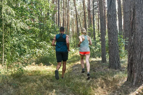 Back view of young sports couple running or jogging in green forest. Beautiful blonde slim woman with tattoo and handsome man wearing sportswear. Concept of modern healthy lifestyle. Sunny summer day